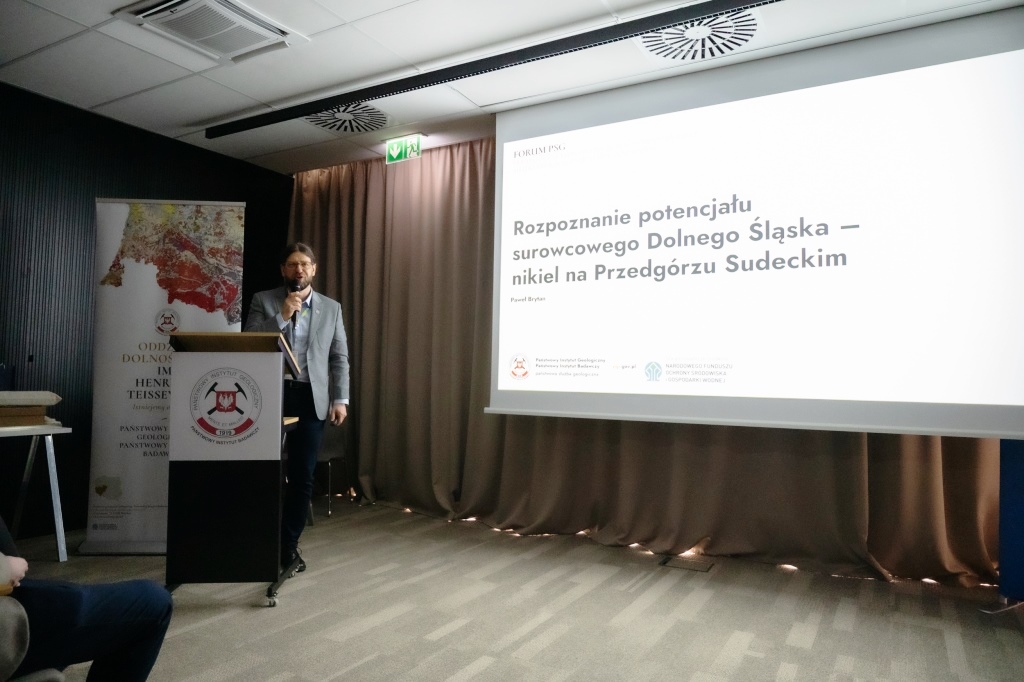 mężczyzna w garniturze przemawia do pełnej sali na tle ekranu z prezentacją